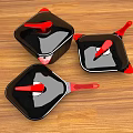 Three Black Cookware Pieces With Red Handles And Lids On Wooden Table