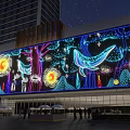 Night View Of Large LED Screen With Colorful Marine Life Sun Trees Tall Buildings Pedestrians