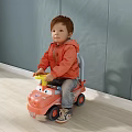 Boy in Orange Jacket Sitting on Yellow Toy Car Against Gray Wall and Light Floor