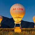 Illuminated AK COM Hot Air Balloons Over Mountain Range And Green Field At Dusk