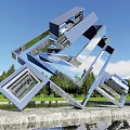 Modern Metallic Landscape Sculpture Featuring Interlocking Geometric Shapes Reflective Surface Green Trees And Blue Sky