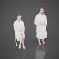 Two Women Wearing White Bathrobes Sitting and Standing on Reflective Floor with Grey Background