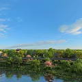 Scenic Park Landscape With Lake Willow Trees Flowering Plants And Traditional Buildings Under Blue Sky