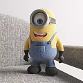 Minion Doll With Large Eye Yellow Body Blue Overalls Black Gloves Standing On Gray Sofa