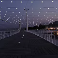 String Lights Hanging Over Wooden Boardwalk With White Railings Bridge And Water Reflection At Evening