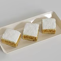 Three square layered pastries with white frosting and golden base on beige tray