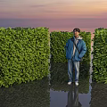 Person Standing Between Three Green Shrub Walls With Sunset Sky And Water Reflection
