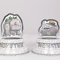 Two Cartoon Monster Figurines On White Speckled Bases With Green Leaf Decor And Hat Accessory