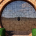 Black Wrought Iron Spider Web Design Gate With Red Brick Arch And Green Shrubs