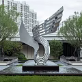 Streamlined Silver Metal Sculpture In Outdoor Space With Water Fountain Trees And High Rise Buildings