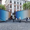 Outdoor Sculpture With Colorful Corrugated Panels In Urban Square With Pedestrians And Historic Buildings