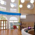 Bright Kindergarten Indoor Space With Round Windows Hanging Lights Wooden Decor And Teacher Child Interaction