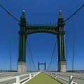 Suspension Bridge With Green Towers Over Calm Water Street Lamps Blue Sky And Reflection