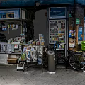 Commercial Street Scene With Newsstand Newspapers Magazines Vending Machine Bicycle And Stone Pavement