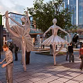 Vibrant Urban Commercial Street with Ballerina Sculptures Pedestrians Modern Buildings and Green Plants