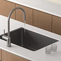 Black Kitchen Sink With Faucet Water Flowing And Glass Cups On White Countertop Wooden Cabinets