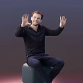 Man Sitting On Green Stool Raising Hands With Palms Out In Black Shirt And Blue Pants