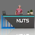 Exhibition Reception Desk Featuring NUTS Logo Blue Frame Gray Countertop Laptop And Potted Plants