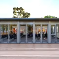 Modern Building Exterior With Large Glass Doors Wooden Deck And Green Trees Under Blue Sky