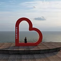Red Heart Shaped Garden Ornament With LOVE LAND Text Overlooking Sea View On Wooden Platform