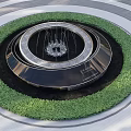 Circular Water Fountain With Multi Tiered Design Surrounded By Green Foliage In Outdoor Plaza