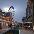 Illuminated Commercial Street With Ferris Wheel Outdoor Seating Pedestrians Flower Beds And Buildings