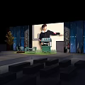 Person Standing On Stage With Blue Beige Walls Green Lawn Area And Small Pavilion Benches