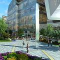 Modern Commercial Street With Glass Facade Buildings Pedestrians Green Plants And Flower Beds