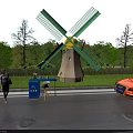 Green Windmill With Yellow Center By Roadside Recycling Bin Orange Sports Car And Trees