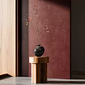 Black Vase With Small Orange Flowers On Wooden Cylindrical Pedestal Against Deep Red Wall