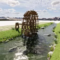 Scenic Waterfront Landscape With Large Wooden Waterwheel By Stream Green Grass And Stones