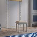 Upholstered Beige Cushion Stool With White Legs Against Gold Nailhead Trim Wall And Wooden Floor