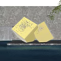 Golden Perforated Cube Sculptures on Waterfront Platform with Waterfall Reflection and Green Plants