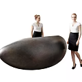 Two Women In White Blouses And Black Skirts Standing By Large Oval Object