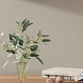 Transparent Vase Holding Green Leaves And White Flowers Against Light Gray Wall With White Book