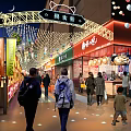 Bustling Commercial Street At Night With Decorative Lights Pedestrians And Colorful Storefronts