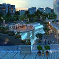 Modern Commercial Street in Evening with Buildings Green Plants Fountains Pedestrians Lighting and Outdoor Seating