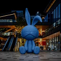 Blue Rabbit Inflatable Model In Outdoor Commercial Plaza At Night With Buildings Escalator And Lighting