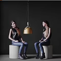 Two women sitting chatting on white cylindrical stools with gold pendant light against dark background