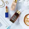 Elegant Cosmetics Display Featuring Lipstick Foundation Facial Serum Gold Necklace Rings White Flowers And Coffee