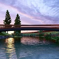 Wooden Bridge Over River With Sunset Sky Purple Pink Clouds And Trees
