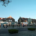 European Style Commercial Street with Red Brick Buildings Pedestrians Outdoor Seating Greenery and Clear Sky