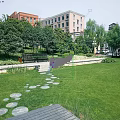 Park Landscape Featuring Green Lawn Circular Stone Stepping Stones Wooden Deck Bench Trees And Buildings