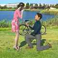 Man Kneeling To Propose To Woman Outdoors By Lake With Bicycle