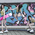 Stylish Women Skateboarding With Pink Skateboard Wearing Yellow Tank Tops Against Colorful Graffiti Wall