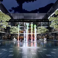 Modern Urban Square With Colorful Fountain Trees Pedestrians And Glass Buildings
