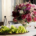 Vibrant Mixed Flowers In Glass Vase With Green Grapes On Tray And Wooden Wall Background