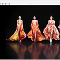Four women in flowing orange and yellow gradient long dresses with reflections on black background