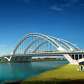 Modern Arch Bridge Over Blue River With Green Grass And People Walking