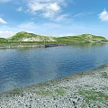 Serene Lakeside Natural Scenery With Calm Blue Water Green Hills White Clouds And Pebbled Shore